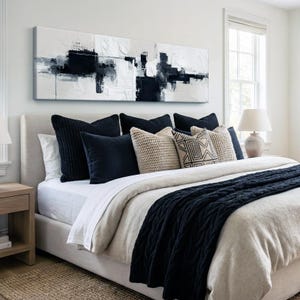 May include: A modern bedroom with a neutral color palette. A large abstract painting in black and white hangs above a bed with navy blue pillows and a matching cable-knit throw. A beige lamp sits on a nightstand.