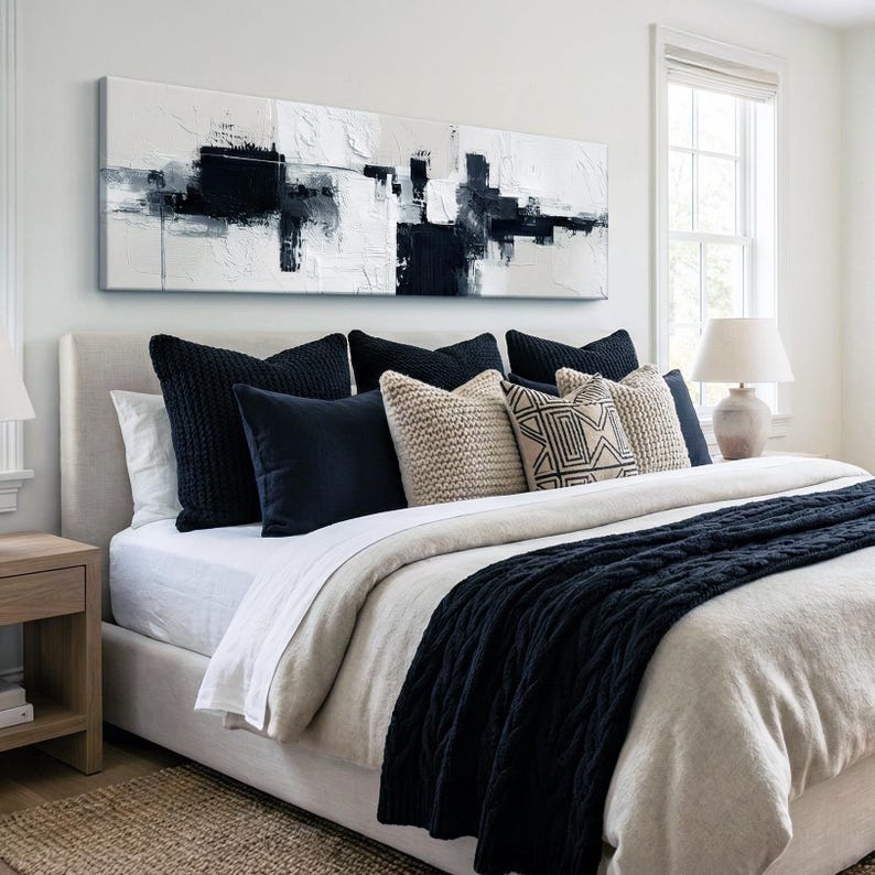 May include: A modern bedroom with a neutral color palette. A large abstract painting in black and white hangs above a bed with navy blue pillows and a matching cable-knit throw. A beige lamp sits on a nightstand.
