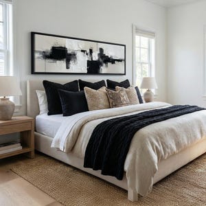 May include: A modern bedroom featuring a bed with a neutral-toned headboard and bedding. Black and beige decorative pillows are arranged on the bed. A black cable-knit throw blanket adds texture. A large abstract painting hangs above the bed.