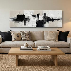 May include: Abstract black, white, and beige painting on canvas, mounted above a beige sofa with black and patterned throw pillows. A wooden coffee table with books and a vase sits in front of the sofa, on a woven rug.