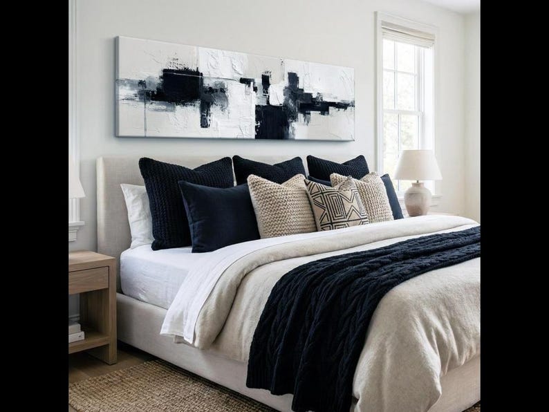 May include: A modern bedroom with a neutral color palette. The bed is made with white linens, navy blue pillows, and a navy blue cable-knit throw. Above the bed is a large abstract painting in black and white.