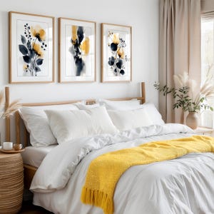 May include: Three framed floral art prints with yellow and navy watercolour designs hang above a bed with white bedding and a yellow throw blanket. The room has a neutral colour palette with natural light.