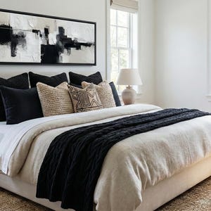 May include: A bedroom with a neutral color palette. The bed is made with a cream-colored comforter and a black knitted throw. Decorative pillows in black, beige, and patterned designs are arranged on the bed. A large abstract painting hangs above the bed.