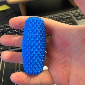 May include: A bright blue, cylindrical object with a textured surface of triangular spikes. The object is held in a hand, showcasing its size and shape. The design suggests a unique, possibly 3D-printed, decorative item.