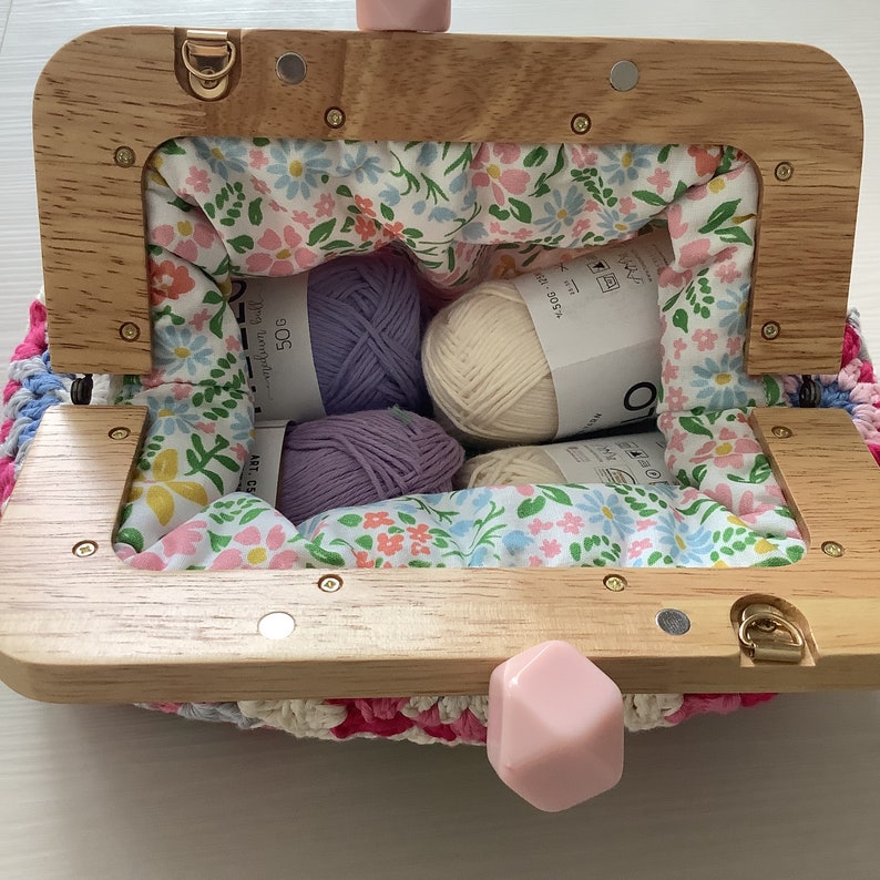 Puede incluir: Una caja de madera con un forro de tela floral y un asa rosa. La caja contiene tres ovillos de lana, dos morados y uno blanco.