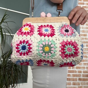 Puede incluir: Un bolso de crochet con un marco de madera y un patr&oacute;n floral rosa y verde. El bolso tiene una correa marr&oacute;n y est&aacute; siendo sostenido por una persona.