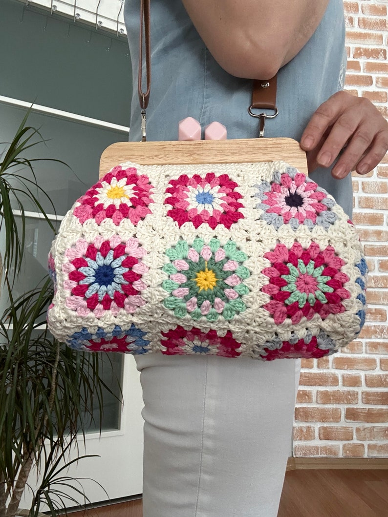 Puede incluir: Un bolso de crochet con un marco de madera y un patr&oacute;n floral rosa y verde. El bolso tiene una correa marr&oacute;n y est&aacute; siendo sostenido por una persona.
