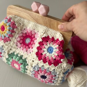 Puede incluir: Un bolso de ganchillo con un marco de madera y cierres de pl&aacute;stico rosa. El bolso est&aacute; hecho de cuadrados de la abuela en varios colores, incluyendo rosa, azul, verde y amarillo.