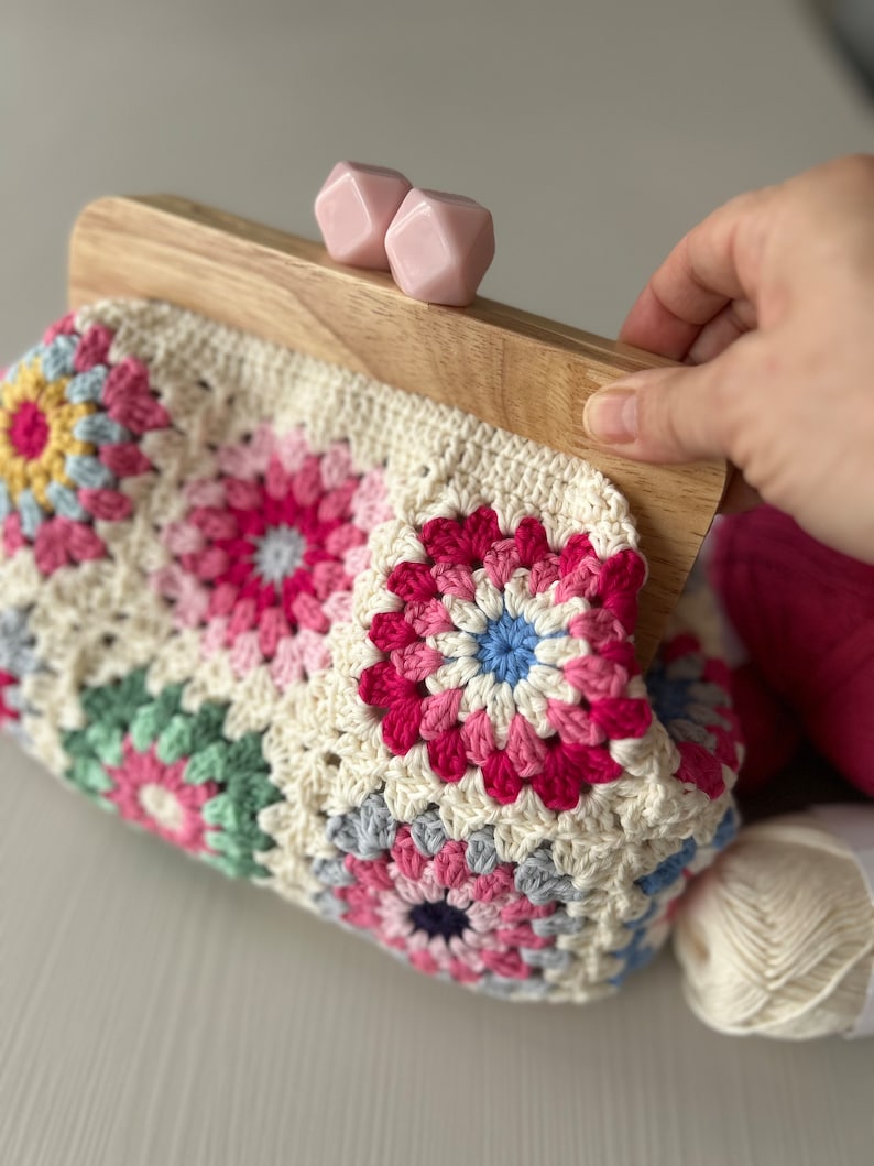 Puede incluir: Un bolso de ganchillo con un marco de madera y cierres de pl&aacute;stico rosa. El bolso est&aacute; hecho de cuadrados de la abuela en varios colores, incluyendo rosa, azul, verde y amarillo.