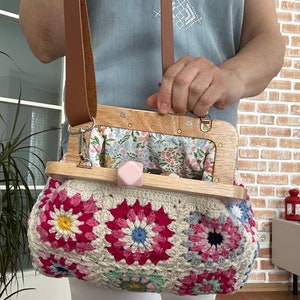 Puede incluir: Un bolso de crochet con un patr&oacute;n floral en tonos de rosa, blanco y azul. El bolso tiene un marco de madera con un forro de tela floral y una correa de cuero marr&oacute;n.