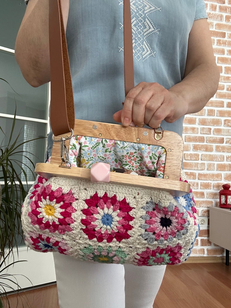 Puede incluir: Un bolso de crochet con un patr&oacute;n floral en tonos de rosa, blanco y azul. El bolso tiene un marco de madera con un forro de tela floral y una correa de cuero marr&oacute;n.