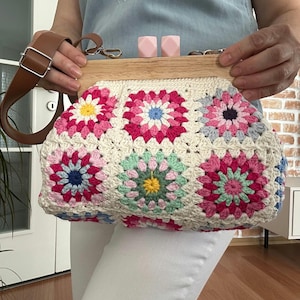 Puede incluir: Un bolso blanco de crochet con un marco de madera y dise&ntilde;os florales rosas, azules y verdes. El bolso tiene una correa de cuero marr&oacute;n.