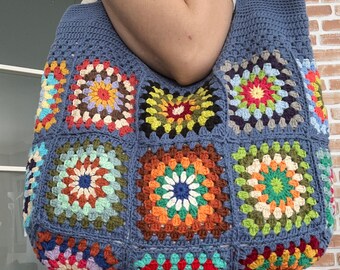 Puede incluir: Una bolsa de mano de crochet azul con diseños de granny squares coloridos. La bolsa tiene una base ancha y plana y dos asas largas.