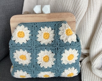 May include: A blue crocheted purse with a wooden frame and white daisy flower designs. The purse has a clasp closure with two white ceramic knobs.