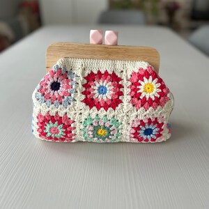 Puede incluir: Un bolso de mano de crochet con un marco de madera y cierres rosas. El bolso est&aacute; hecho de hilo blanco con cuadrados de granny coloridos con flores rosas, rojas, azules, verdes y amarillas.