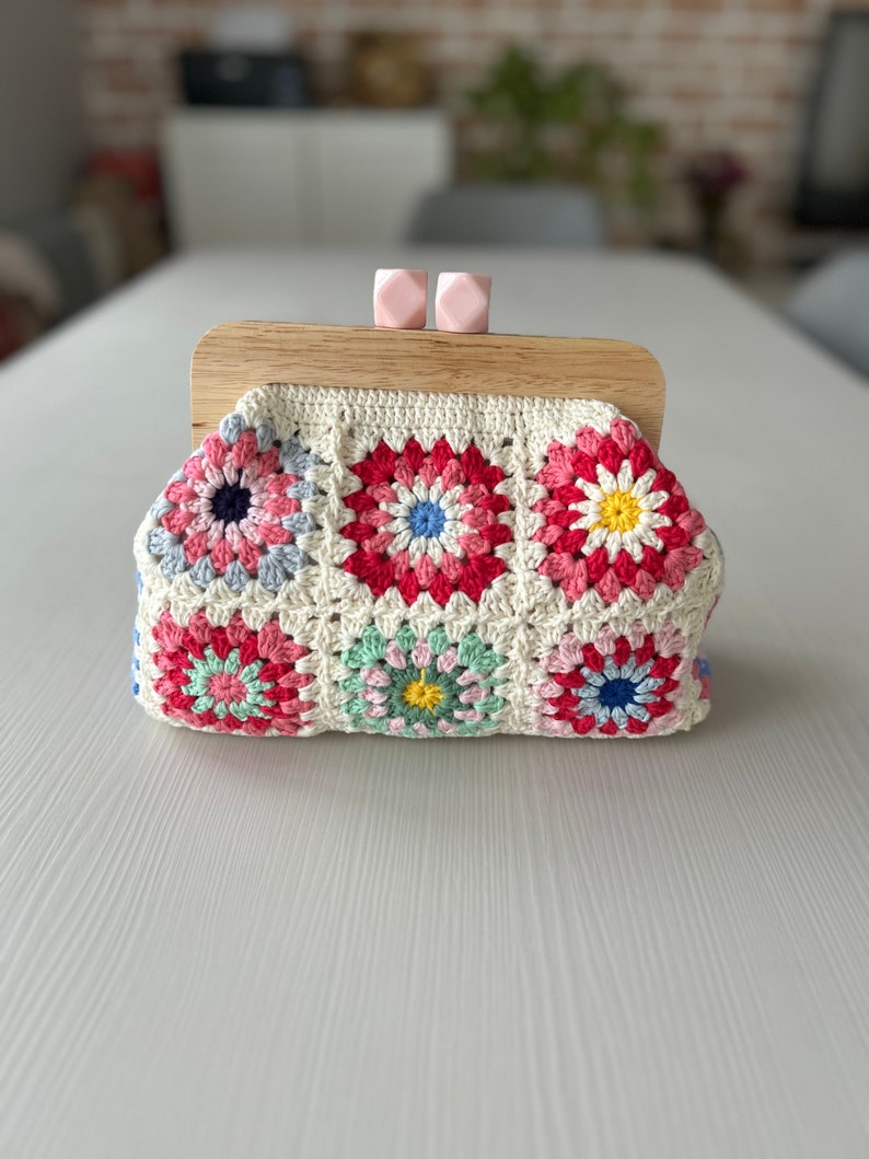 Puede incluir: Un bolso de mano de crochet con un marco de madera y cierres rosas. El bolso est&aacute; hecho de hilo blanco con cuadrados de granny coloridos con flores rosas, rojas, azules, verdes y amarillas.