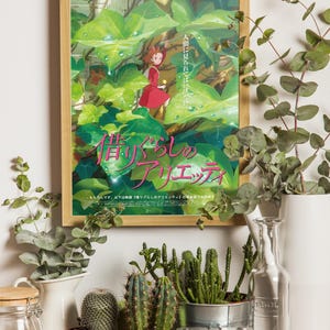 Pu&ograve; includere: Una locandina cinematografica incorniciata di Arrietty - Il mondo segreto sotto il pavimento con un personaggio in abito rosso tra grandi foglie verdi. Il poster include testo giapponese ed &egrave; esposto accanto a cactus, una fotocamera vintage e vasi.