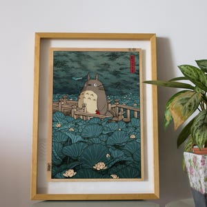 May include: Framed print depicting Totoro in a tranquil aquatic scene. The artwork shows a large, grey Totoro character surrounded by vibrant, teal lily pads and delicate white lotus flowers. The print is encased in a light brown wooden frame.