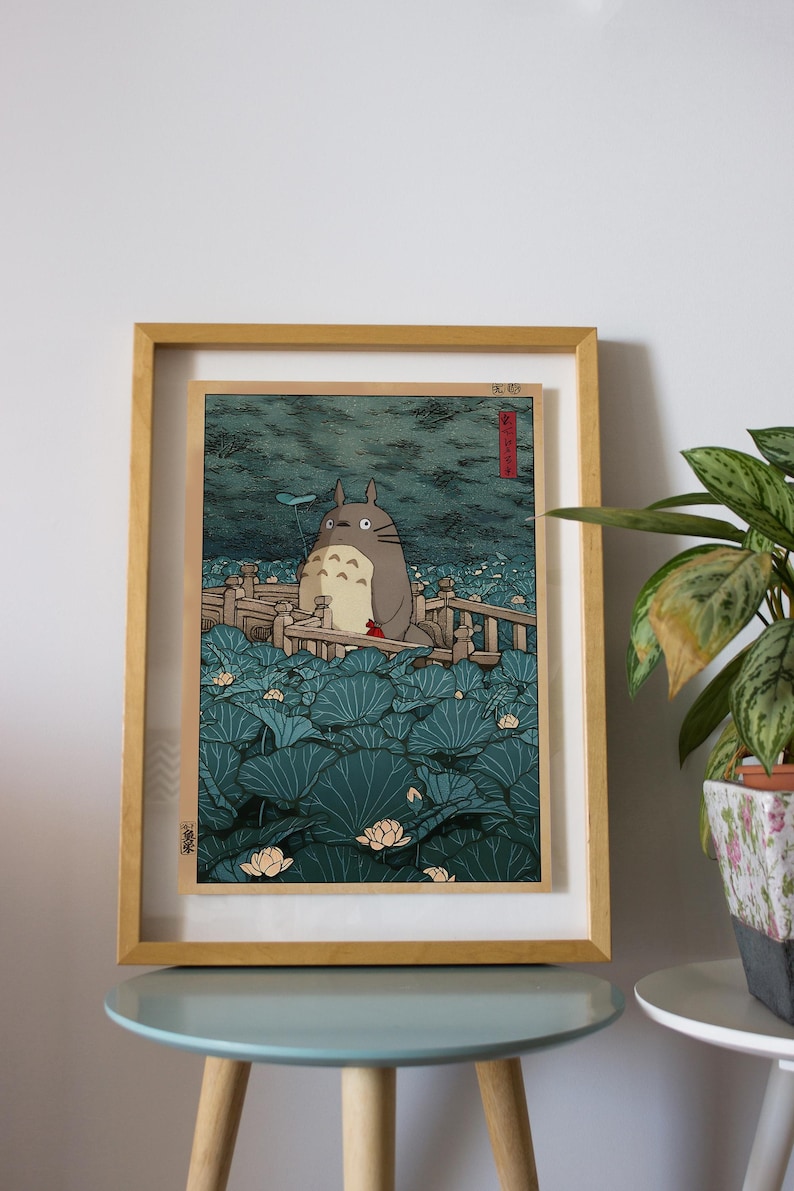 May include: Framed print depicting Totoro in a tranquil aquatic scene. The artwork shows a large, grey Totoro character surrounded by vibrant, teal lily pads and delicate white lotus flowers. The print is encased in a light brown wooden frame.