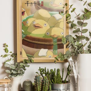 May include: A framed poster with a cartoon image of yellow ducklings in a wooden tub. The poster is hanging on a white wall with green plants and a camera in the foreground.
