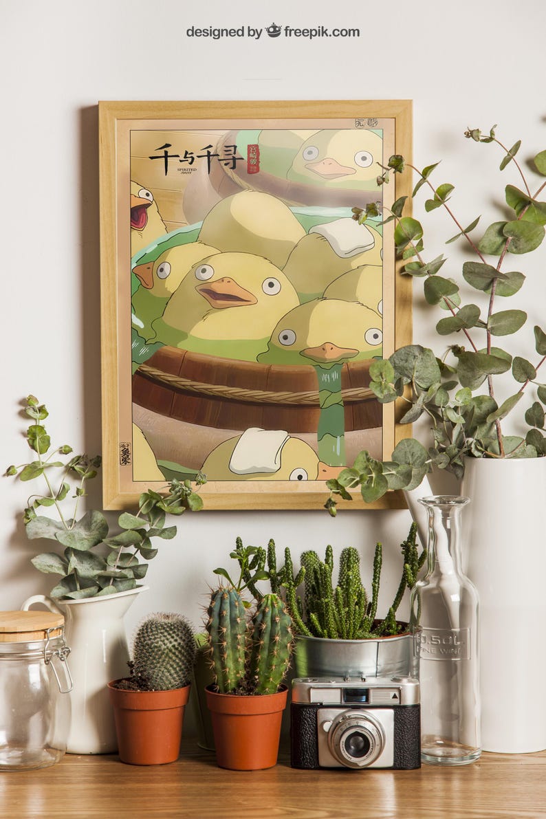May include: A framed poster with a cartoon image of yellow ducklings in a wooden tub. The poster is hanging on a white wall with green plants and a camera in the foreground.