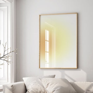 May include: A framed abstract art print with a yellow to white gradient. The print is hanging on a white wall above a white couch with white pillows.