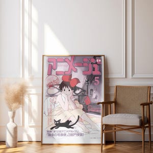 May include: A framed poster featuring Japanese anime artwork. The poster has a pink and white color scheme with Japanese text and an illustration of a girl with a black cat. The poster is leaning against a white wall next to a wooden chair.