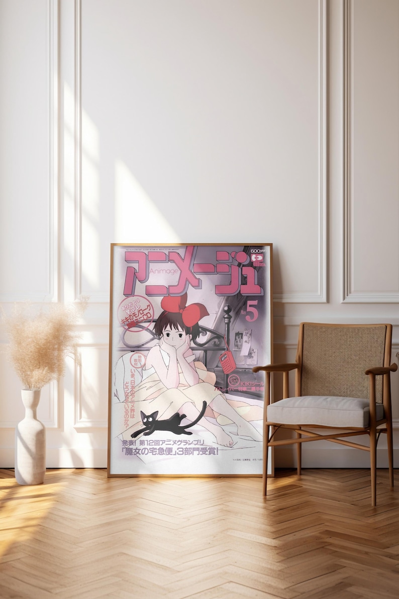 May include: A framed poster featuring Japanese anime artwork. The poster has a pink and white color scheme with Japanese text and an illustration of a girl with a black cat. The poster is leaning against a white wall next to a wooden chair.