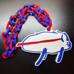 May include: A white, blue, and red Buffalo Bills logo pendant with a chain of red and blue links. The pendant features a white buffalo with blue outlines and a red stripe across the middle. The chain is made of alternating red and blue links.