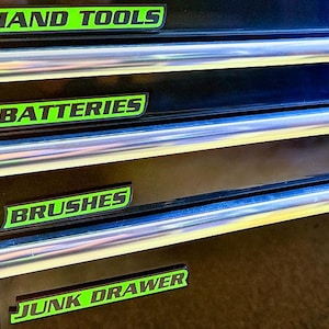 May include: A black tool chest with four drawers, each labeled with green and white text. The labels read "HAND TOOLS," "BATTERIES," "BRUSHES," and "JUNK DRAWER." The drawers have silver trim.