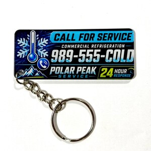May include: A rectangular keychain featuring a blue and black design with a thermometer and snowflake graphic. The text reads CALL FOR SERVICE, COMMERCIAL REFRIGERATION, 989-555-COLD, POLAR PEAK SERVICE, and 24 HOUR RESPONSE with a phone icon.