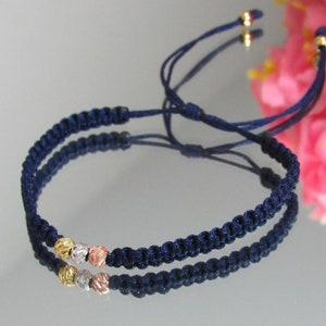 May include: Navy blue macrame bracelet with three textured beads in gold, silver, and rose gold. The bracelet has adjustable sliding knots and gold-colored end caps. This handmade jewelry piece is a stylish accessory.