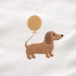 Könnte beinhalten: Gestickter Dackel mit gelbem Ballon auf weißem Hintergrund. Der Hund ist braun mit einem hellbraunen Ohr und einer schwarzen Nase. Der Ballon ist rund und ebenfalls gelb. Die Stickerei hat ein strukturiertes Aussehen.