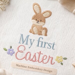 Könnte beinhalten: Bestickter weißer Stoff mit einem braunen Hasen und dem Text "My first Easter" in Blau und Rosa. Blumige Akzente in Lila und Gelb umgeben den Text. Darunter steht "Machine Embroidery Design".