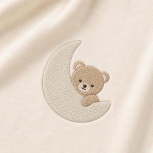 May include: Cream-colored fabric with an embroidered design of a teddy bear nestled in a crescent moon. The bear is light brown with dark brown eyes and nose. The moon is a lighter cream color, and the stitching is visible.