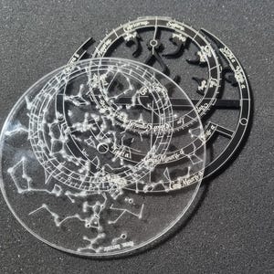 May include: Clear acrylic celestial chart with white etched constellations, zodiac signs, and text. A black, circular frame with zodiac symbols and text is layered on top. The design features intricate details of the night sky.