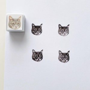 May include: Five rubber stamps featuring a cat's face. The stamps are white with black ink. The cat's face is looking directly at the viewer. The stamps are arranged in a grid pattern.