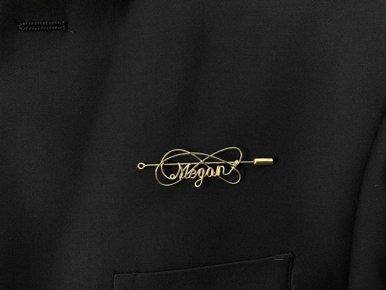 May include: Gold wire lapel pin with the name "Megan" written in cursive.