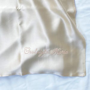 Könnte beinhalten: Elfenbeinfarbener Satin-Kissenbezug mit dem gestickten Text "Goodnight Marie" in zarter Schreibschrift. Der Kissenbezug ist teilweise sichtbar, mit weichen Falten und dezentem Glanz, der auf einer hellblauen Oberfläche liegt.