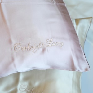 Peut inclure: Gros plan sur deux taies d'oreiller en soie. La taie d'oreiller supérieure est rose clair avec le texte brodé "Goodnight Lacey" en écriture cursive. La taie d'oreiller inférieure est de couleur crème avec le texte brodé "Goodnight".