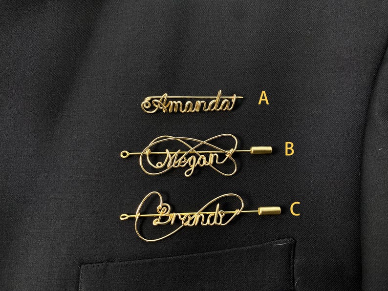 May include: Three gold wire name pins on a black fabric background. The pins are shaped like infinity symbols and have the names "Amanda", "Megan", and "Brenda" written on them.
