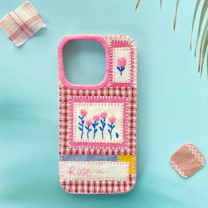 May include: A patchwork phone case in pink and white, adorned with embroidered flowers. The case has a gingham pattern, a pink border, and the word "Rose" embroidered on the front. It's made from small fabric patches, with a stitched design.
