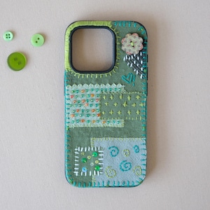 May include: A rectangular olive green phone case featuring decorative fabric patches, hand-stitched embroidery, colorful beads, and a floral button. Three loose green buttons are positioned to the left of the phone case.