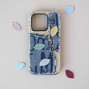 May include: A rectangular phone case featuring a patchwork design of dark blue denim fabric with white lettering and beige textile. The case is decorated with small, embroidered leaf-shaped appliques in yellow, green, light blue, and grey colors.