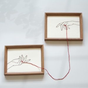 May include: Two framed art pieces with a minimalist design. Each frame is a warm brown, housing a cream-colored background. Black line art depicts hands interacting with a red thread. The thread connects the two pieces, creating a visual narrative.