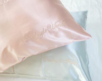 May include: Two satin pillowcases, one light pink and one light blue, are stacked. The pink pillowcase has "Goodnight Wenda" embroidered in a cursive font. The blue pillowcase has "Goodnight Trana" embroidered in a similar style. Both pillowcases have a shiny finish.