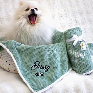 May include: A white Pomeranian dog yawns in a basket with a green blanket and matching rolled-up towel. The blanket is embroidered with the name "Daisy" and paw prints. The towel is labeled "Angel" with a white ribbon and tag.