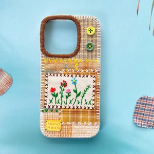 May include: A handcrafted phone case with a patchwork design and floral embroidery. The case has a brown camera aperture, yellow and green buttons, and a label reading "Hand Made". The case is made from various fabrics in shades of brown and yellow.