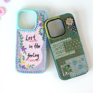 May include: Two phone cases with embroidered fabric covers. The left case is light purple with the text "Lost in the feeling" in black thread, surrounded by floral embroidery. The right case is olive green with patchwork stitching and a button.