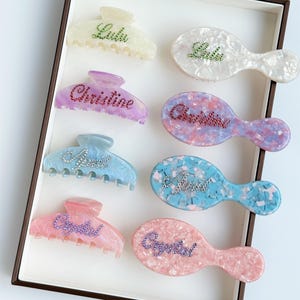 May include: Assortment of eight hair clips and hairbrushes in various colors, including white, pink, purple, and blue. Each clip and brush features a different name, including Lulu, Christine, Amy, and Crystal, spelled out in rhinestones.